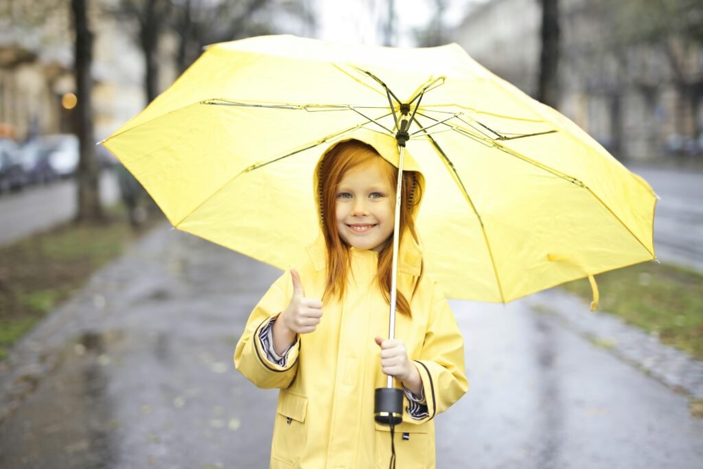 Jeune fille souriant sous un parapluie jaune dans une rue pluvieuse, montrant un pouce levé.
